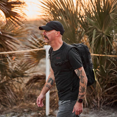 Man in GORUCK Subtle Spearhead Tee and cap rucking with backpack outdoors at sunset