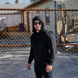 Man wearing black GORUCK hoodie and backpack, standing outdoors by a chain link fence