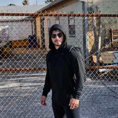 Man wearing black GORUCK hoodie and backpack, standing outdoors by a chain link fence