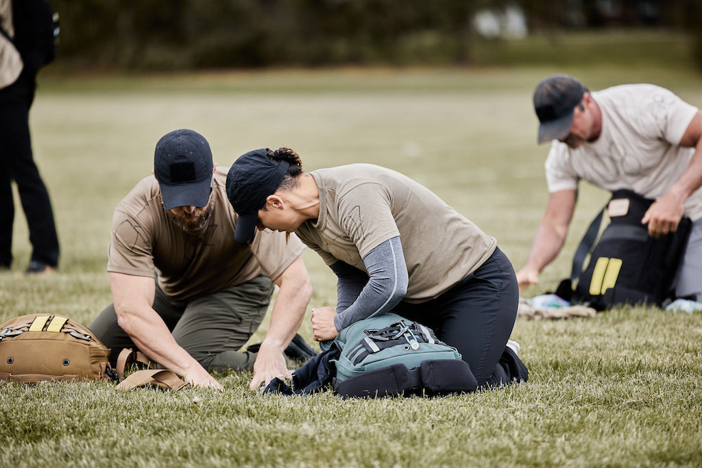 Two people kneeling on grass, packing backpacks, with another person doing the same in the background.