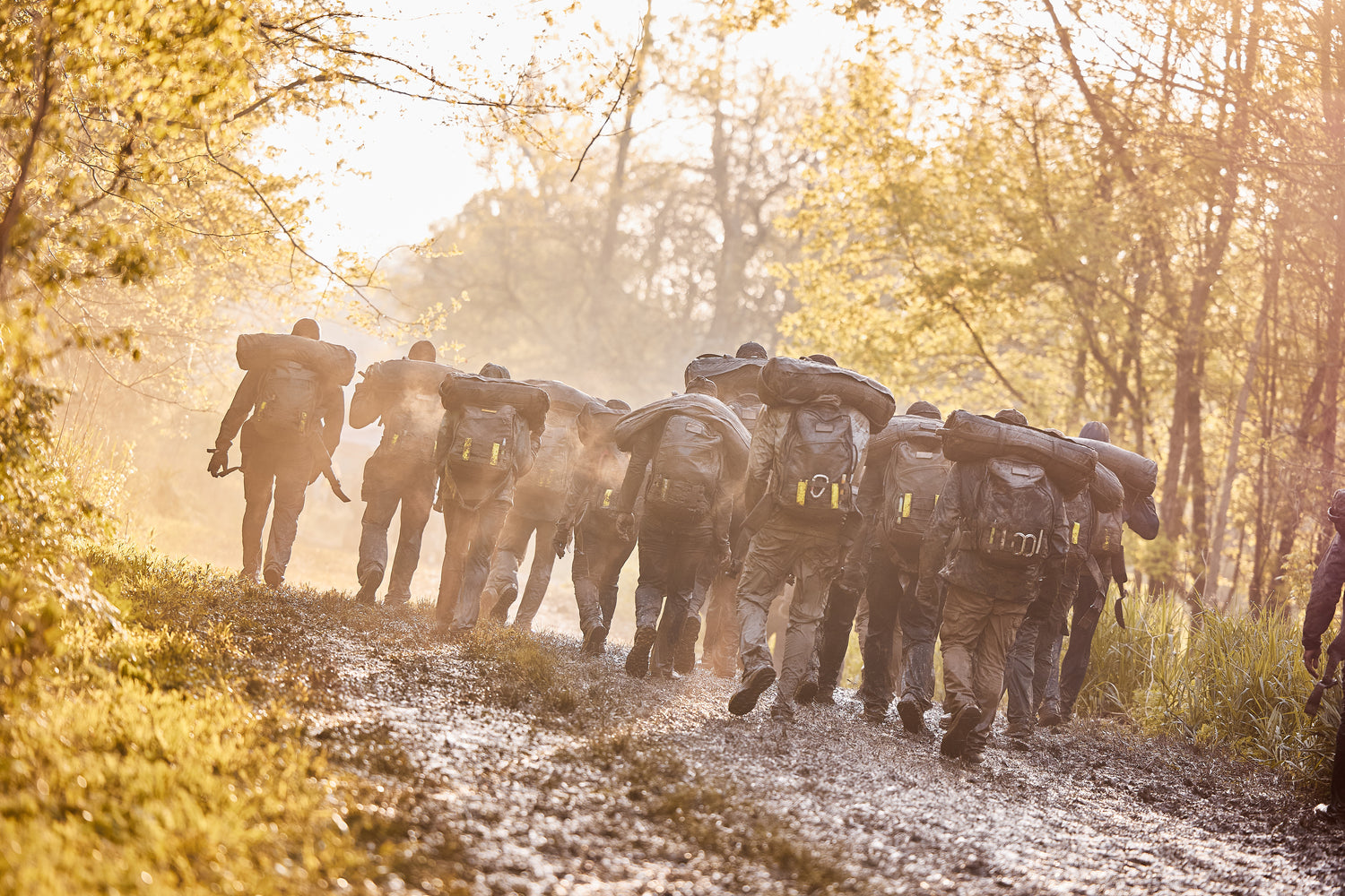 A group of people carrying heavy backpacks walks up a muddy path in a forest at sunrise.