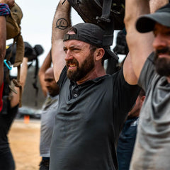 Group of men in GORUCK hats and gear lifting rucksacks overhead during outdoor rucking event