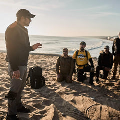 GORUCK rucking group training on sandy beach near ocean, wearing outdoor tactical gear