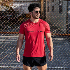 A man in sunglasses and a red Men’s Performance Tee - ToughMesh stands before a chain-link fence on a sunny day, ready to take on the heat with the shirt’s quick-drying performance.