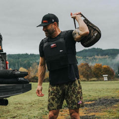 A man in a black cap, black shirt, and camouflage shorts is working out in the open air, sporting the GORUCK Training Weight Vest 2.0. Amidst a grassy field with trees around him, he lifts a sandbag over his shoulder. A truck parked nearby emphasizes his commitment to rigorous training with the adjustable GORUCK vest as part of his routine.