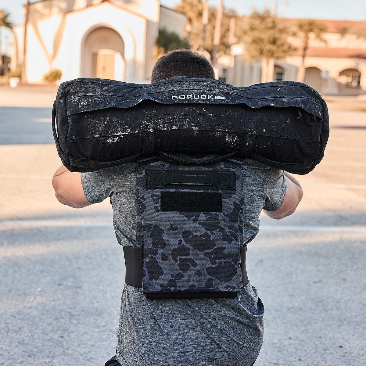 A man outdoors, facing away from the camera, carries a heavy sandbag on his shoulders while wearing the Training Weight Vest 2.0.