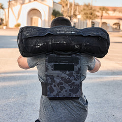 A man outdoors, facing away from the camera, carries a heavy sandbag on his shoulders while wearing the Training Weight Vest 2.0.