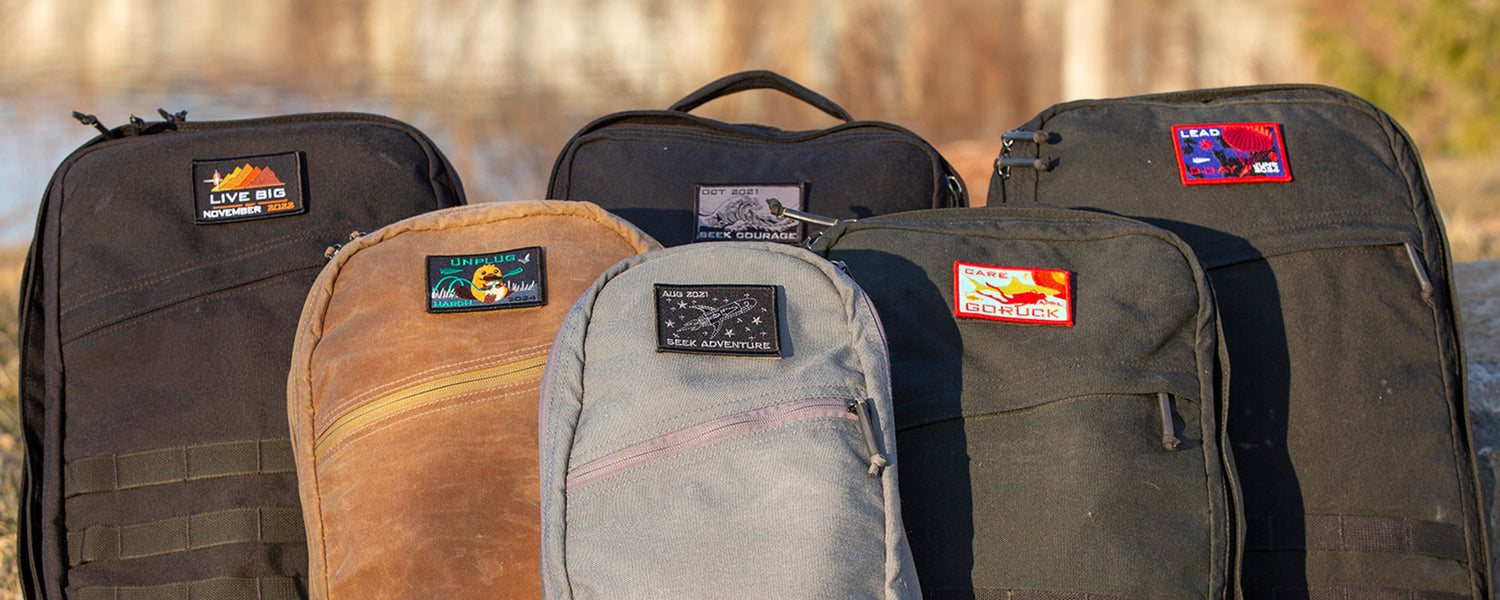 Six backpacks with colorful patches are lined up outdoors in sunlight, with blurred nature in the background.