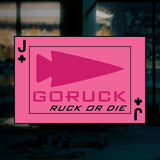 The "RUCK OR DIE" playing card features a bold pink design with an arrowhead, the phrase "GORUCK RUCK OR DIE," and a Jack of clubs symbol—ideal for rucking enthusiasts and dedicated members of the goruck.mommunity, showcased against a blurred room backdrop.
