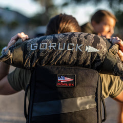 Person rucking outdoors with GORUCK sandbag and rucksack featuring American flag patch