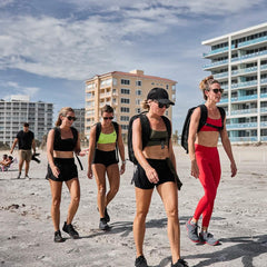 Four women wearing athletic gear and GORUCK training backpacks walking on a sandy beach with buildings in the background.