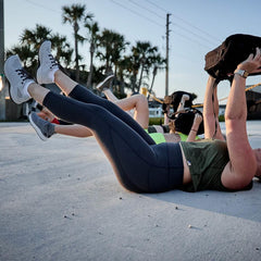 Women wearing GORUCK Ballistic Trainers exercising outdoors on pavement, raising rucksacks, palm trees in background.