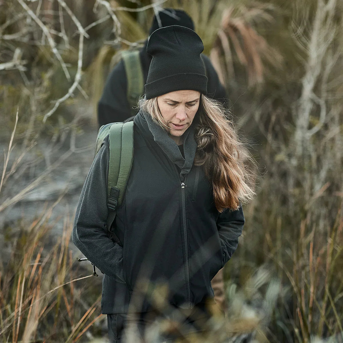 Woman rucking outdoors in black fleece vest, black beanie, and green backpack by GORUCK