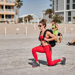 Woman in red GORUCK ToughFlex training leggings lunging on the beach with rucksack