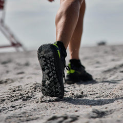 A person walks on a sandy beach, their legs and the sole of their Midnight Frogskin + Acid Lime Women's Rough Runner lightweight running shoes by GORUCK captured in focus. The sky is cloudy, and the sand appears coarse beneath the GRADIENT DENSITY EVA MIDSOLE.