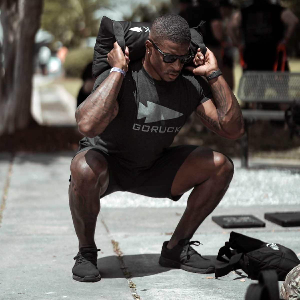 Man doing outdoor squats with GORUCK sandbag, wearing GORUCK shirt and sunglasses