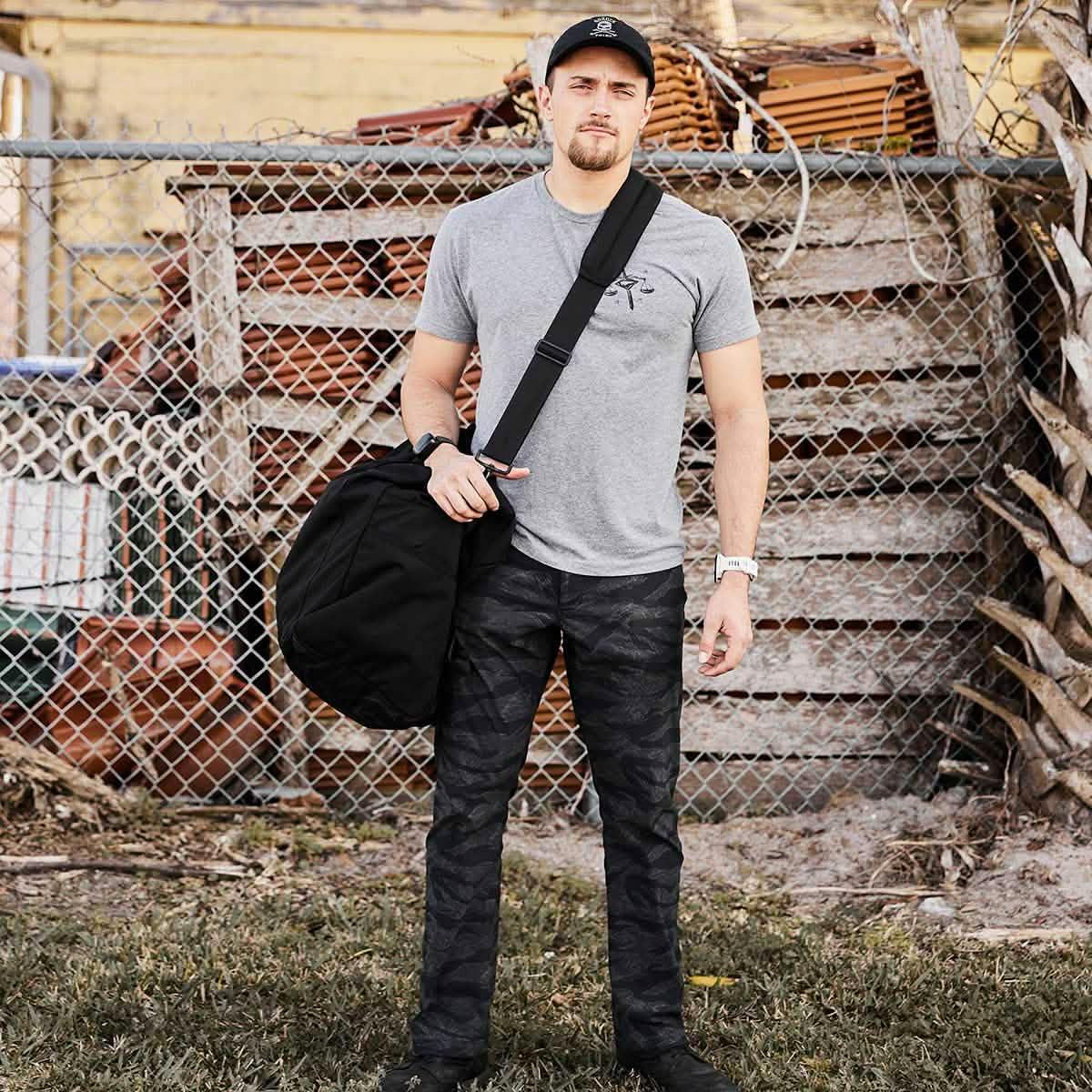 Man in GORUCK gear with rucksack standing outdoors by fence, wearing gray shirt and camo pants