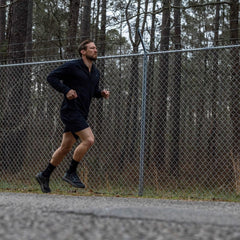 Man running outdoors wearing GORUCK gear by a chain-link fence and pine trees