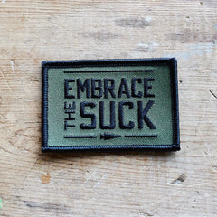 GORUCK green embroidered morale patch with 'Embrace the Suck' text on wood surface