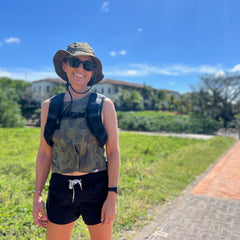 Smiling woman outdoors on a sunny day, wearing sunglasses and the Boonie Hat - Slick - ToughDry, stands on a path with greenery behind her, enjoying the comfort of its durable ToughDry fabric.