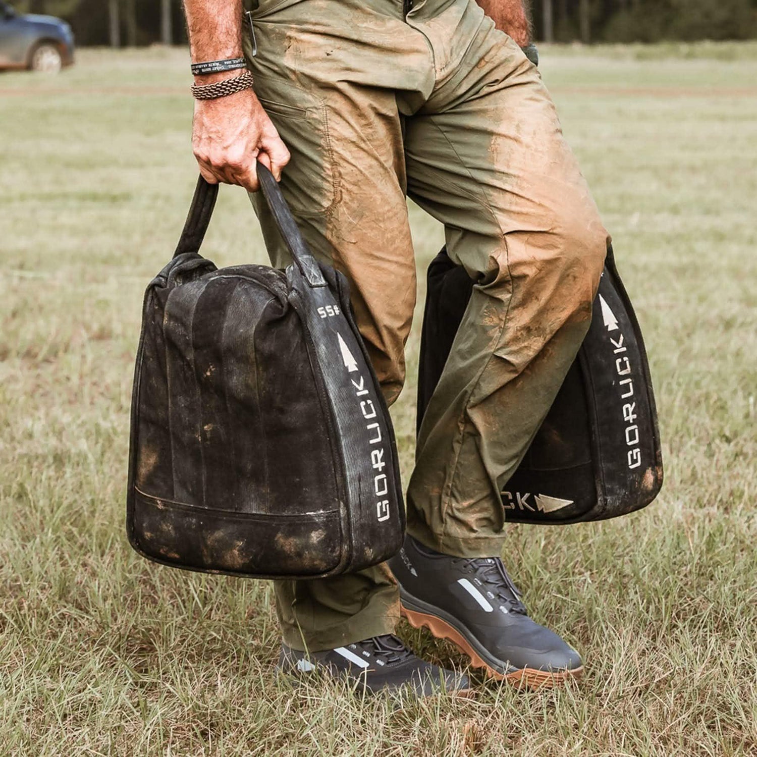 Person carrying two GORUCK sandbags on grass, wearing muddy pants and outdoor shoes