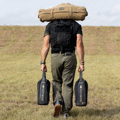 Man wearing a tactical vest carries GORUCK sandbags and gear outdoors on grass field