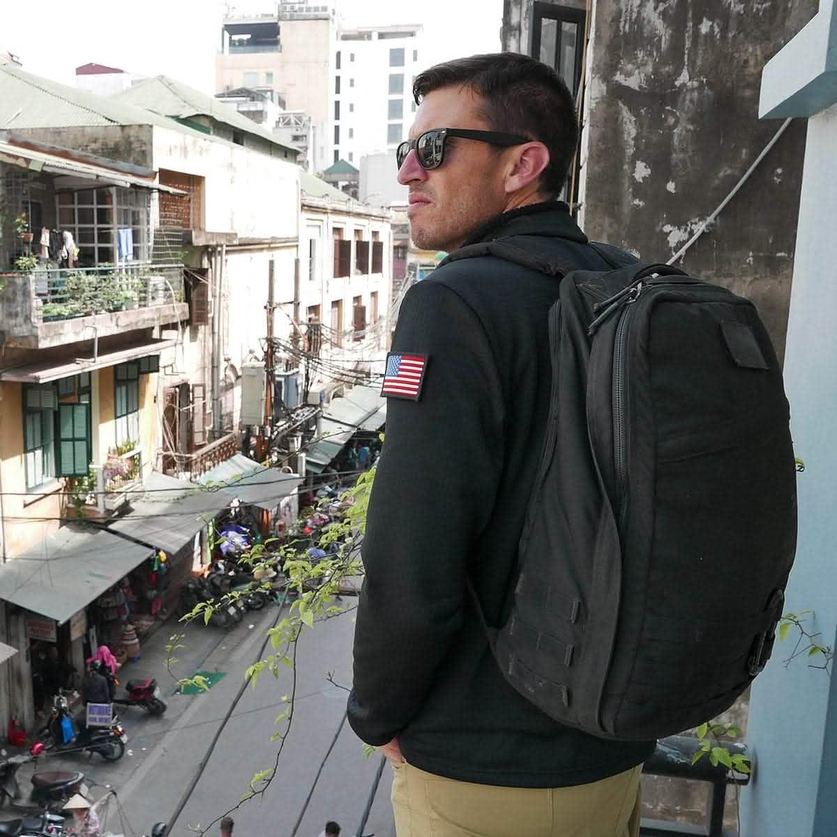 Man wearing GORUCK backpack and jacket with American flag patch, overlooking city street