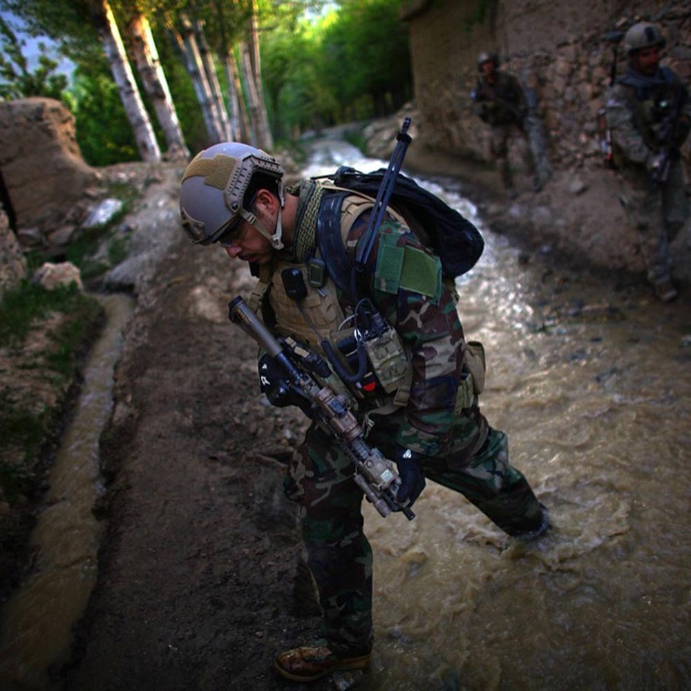 A soldier in camouflage gear crosses a muddy stream, with other soldiers in the background.