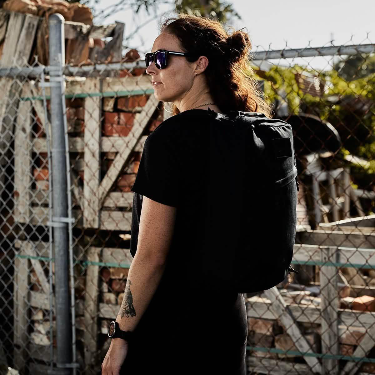 Woman wearing GORUCK black rucksack outdoors by a chain link fence, rucking gear