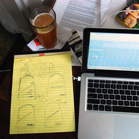 A laptop, beer, and yellow notepad with sketches and notes on a cluttered desk.