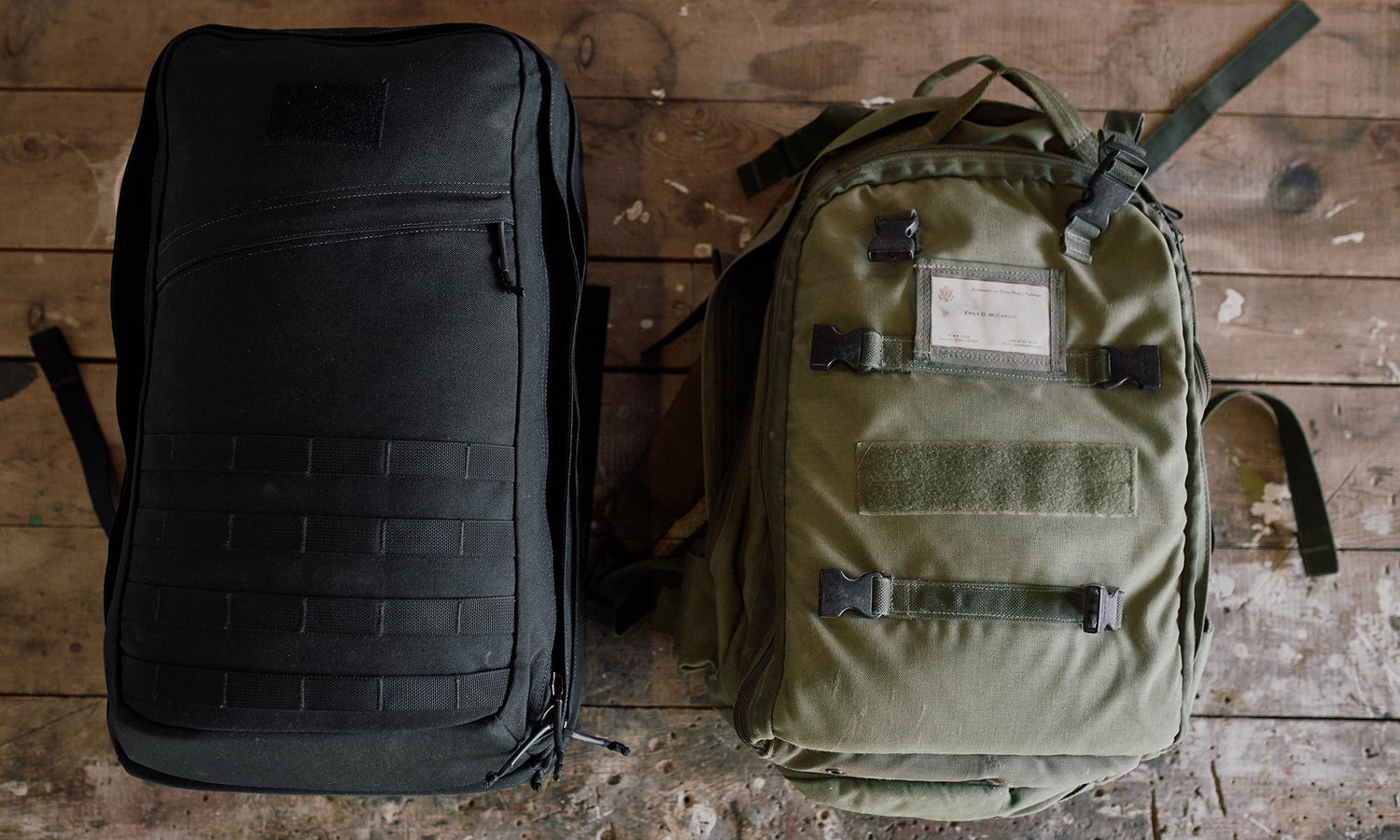 Two backpacks, one black and one green, are placed side by side on a worn wooden floor.
