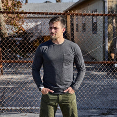 A man wearing the Men's Long Sleeve Pocket Tee - Tri-BlendX in gray stands in front of a chain-link fence on a sunny day.