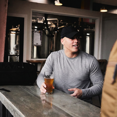 A man in a Men's Slick Long Sleeve - Tri-BlendX shirt and black cap sits at a bar, holding a glass of beer, smiling and chatting.