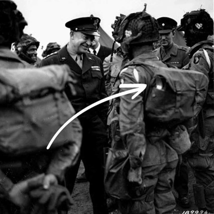 A military officer smiles while talking to paratroopers; an arrow points to a soldier’s gear.