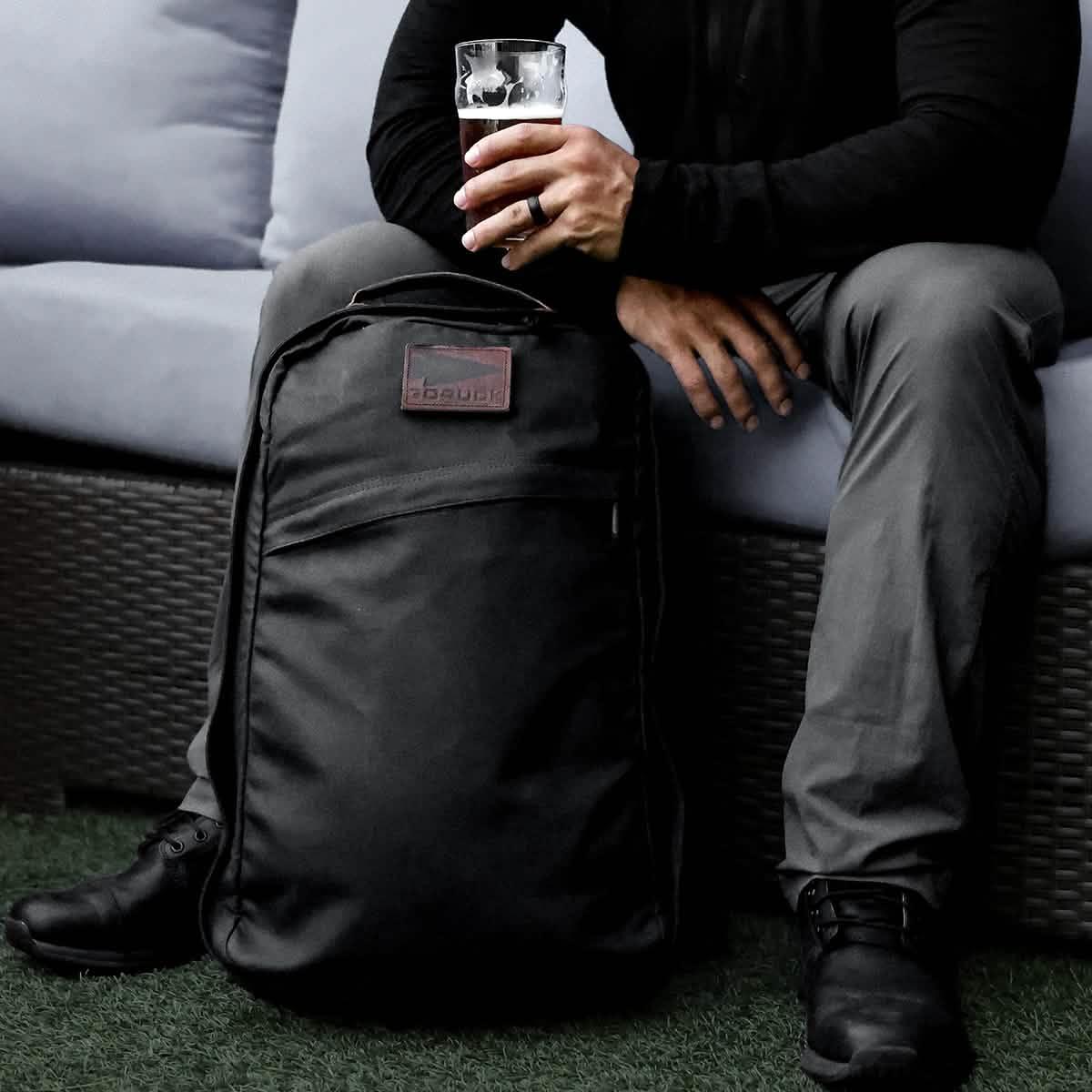 GORUCK black rucksack on grass beside seated man holding a drink, rugged outdoor gear