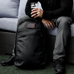 GORUCK black rucksack on grass beside seated man holding a drink, rugged outdoor gear