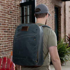 Man wearing GORUCK heritage slate rucksack outdoors, brick wall and plants in background