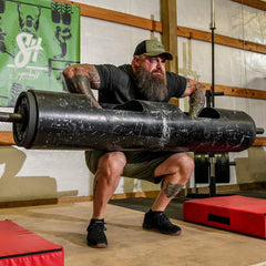 Bearded man squatting with large log barbell in gym, showcasing GORUCK strength training gear.