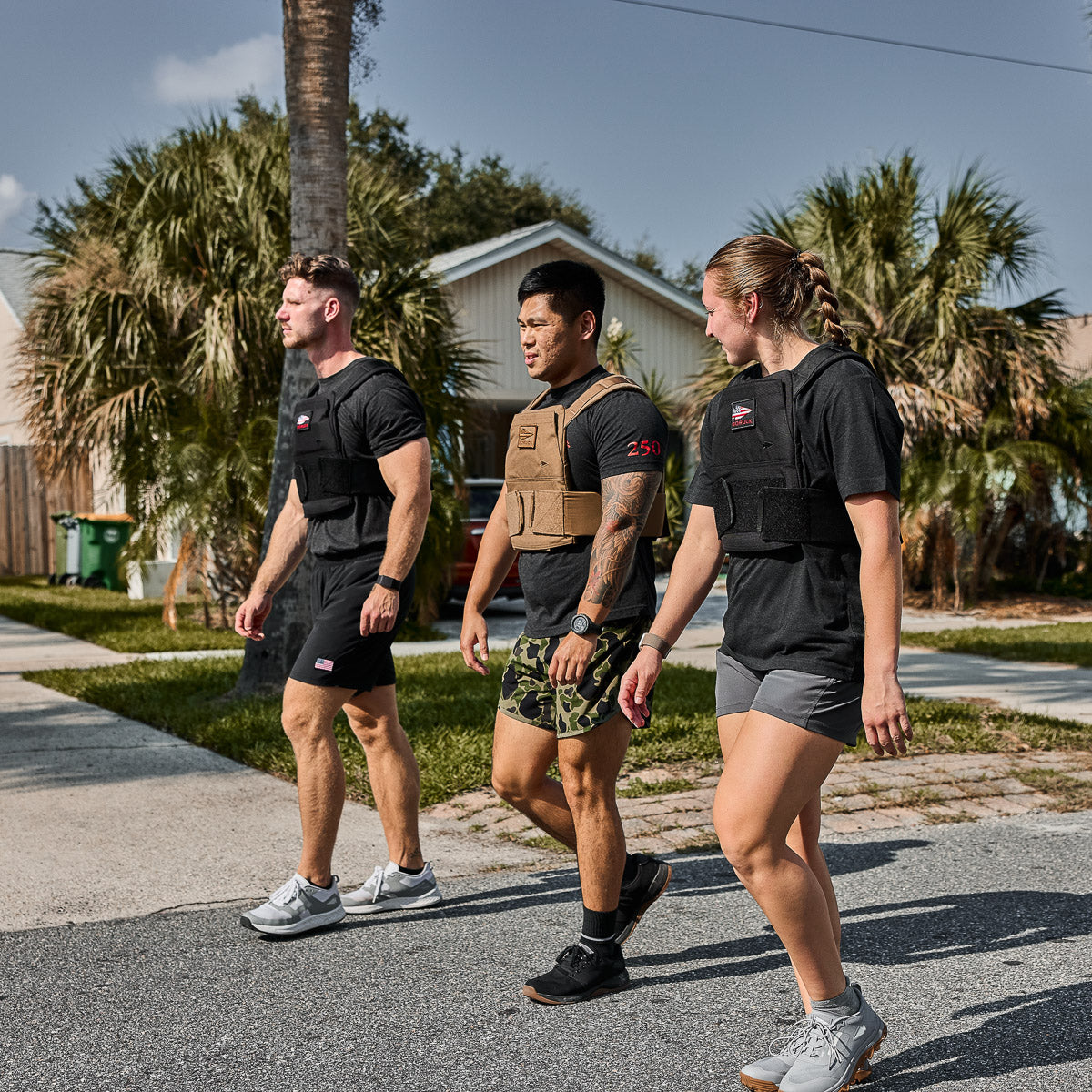 Three people in athletic wear and weighted vests walking together outside on a sunny day near palm trees.