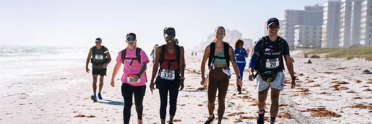 A group of marathon runners smiles while walking along a sandy beach with buildings in the background.