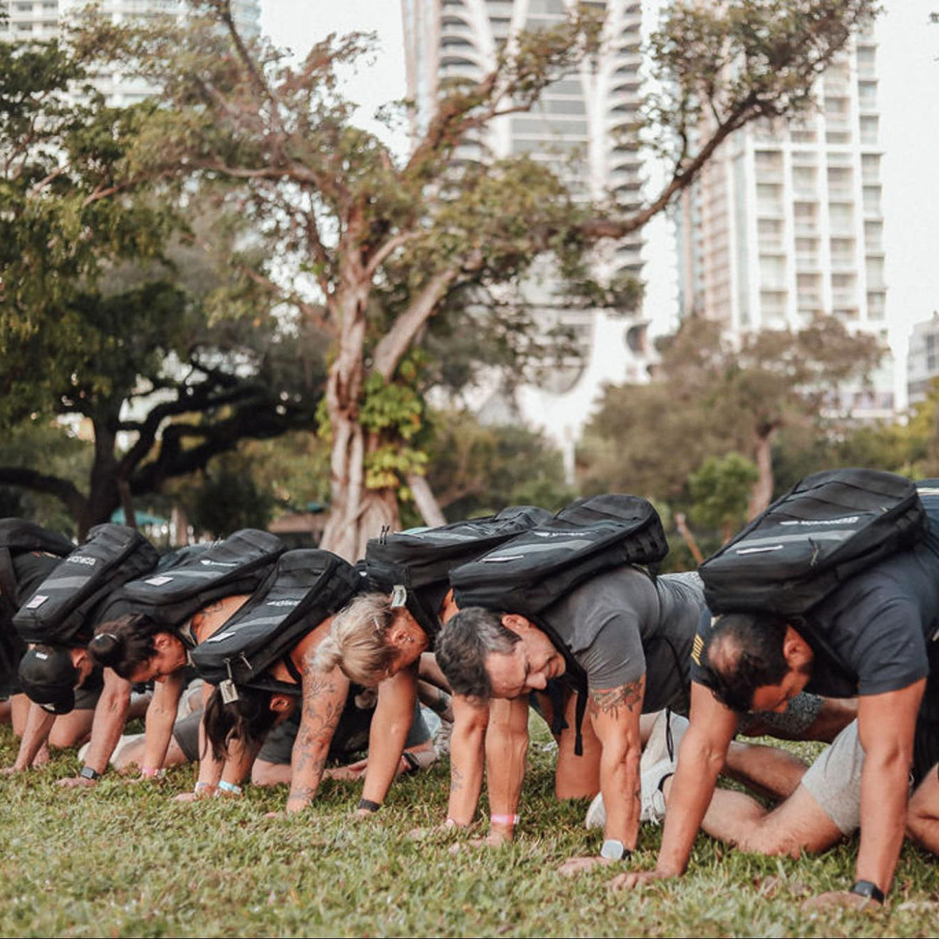 Six people wearing backpacks doing crawling exercises on grass in an outdoor urban park setting.