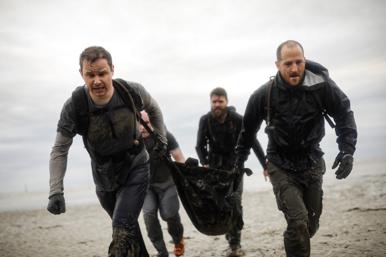 Four men in muddy clothes run on a beach, carrying a heavy object together with determined expressions.