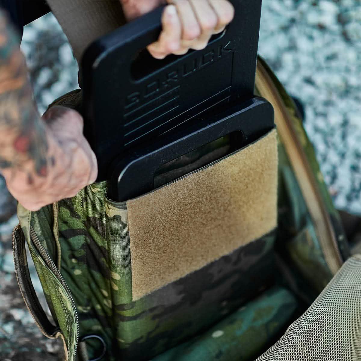 Person placing a GORUCK weight plate into a camo rucking backpack outdoors
