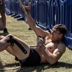 Athlete in shorts struggles during GORUCK outdoor training, gripping rope on grass field