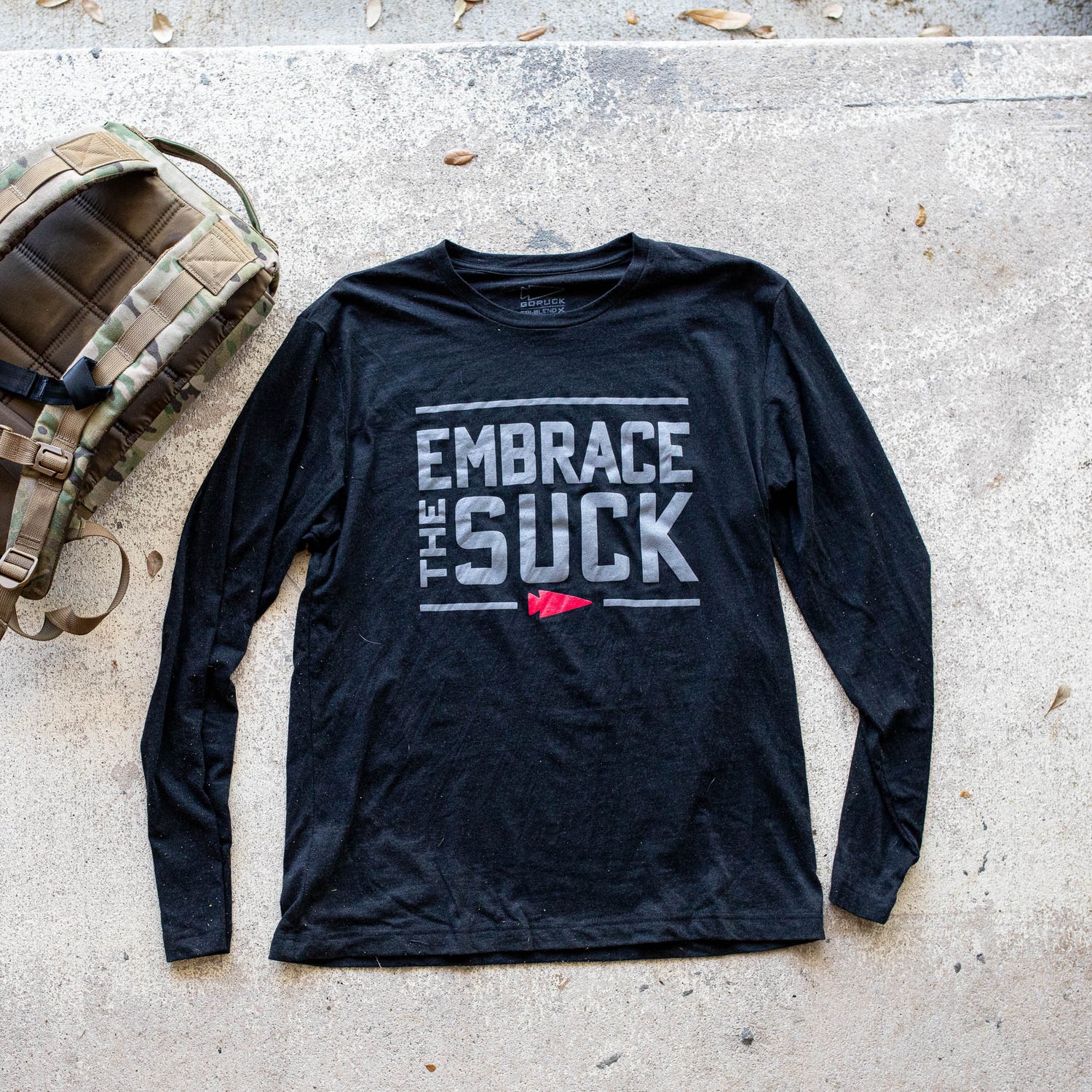 The Men's Embrace the Suck Long Sleeve - Tri-BlendX in black is showcased on concrete beside a camouflage backpack, featuring the 