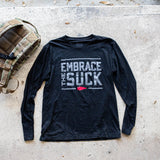 The Men's Embrace the Suck Long Sleeve - Tri-BlendX in black is showcased on concrete beside a camouflage backpack, featuring the "Embrace the Suck" text in premium tri-blend fabric.