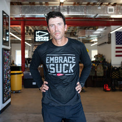 A man in athletic gear stands indoors, hands on hips, wearing the Men's Embrace the Suck Long Sleeve - Tri-BlendX. The premium tri-blend fabric and antimicrobial treatment keep him comfortable and fresh during workouts.