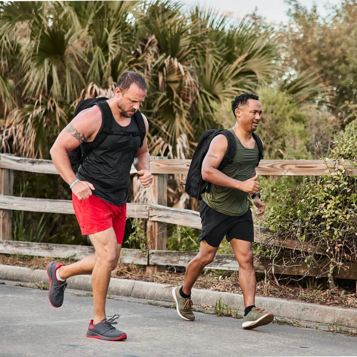 Two men rucking outdoors wearing GORUCK gear, running by a wooden fence with backpacks.