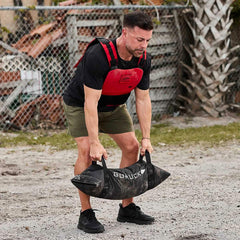 Man wearing GORUCK gear lifting sandbag outdoors, showcasing rucking fitness equipment