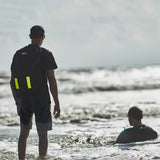 Person with GORUCK backpack in water, rucking on the beach, another person sitting in surf